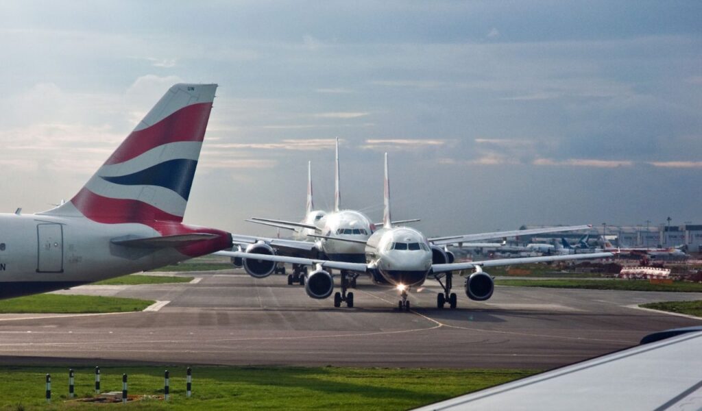 Multiple British Airways planes taxi in line on the runway at Heathrow Airport, with cloudy sky in the background.